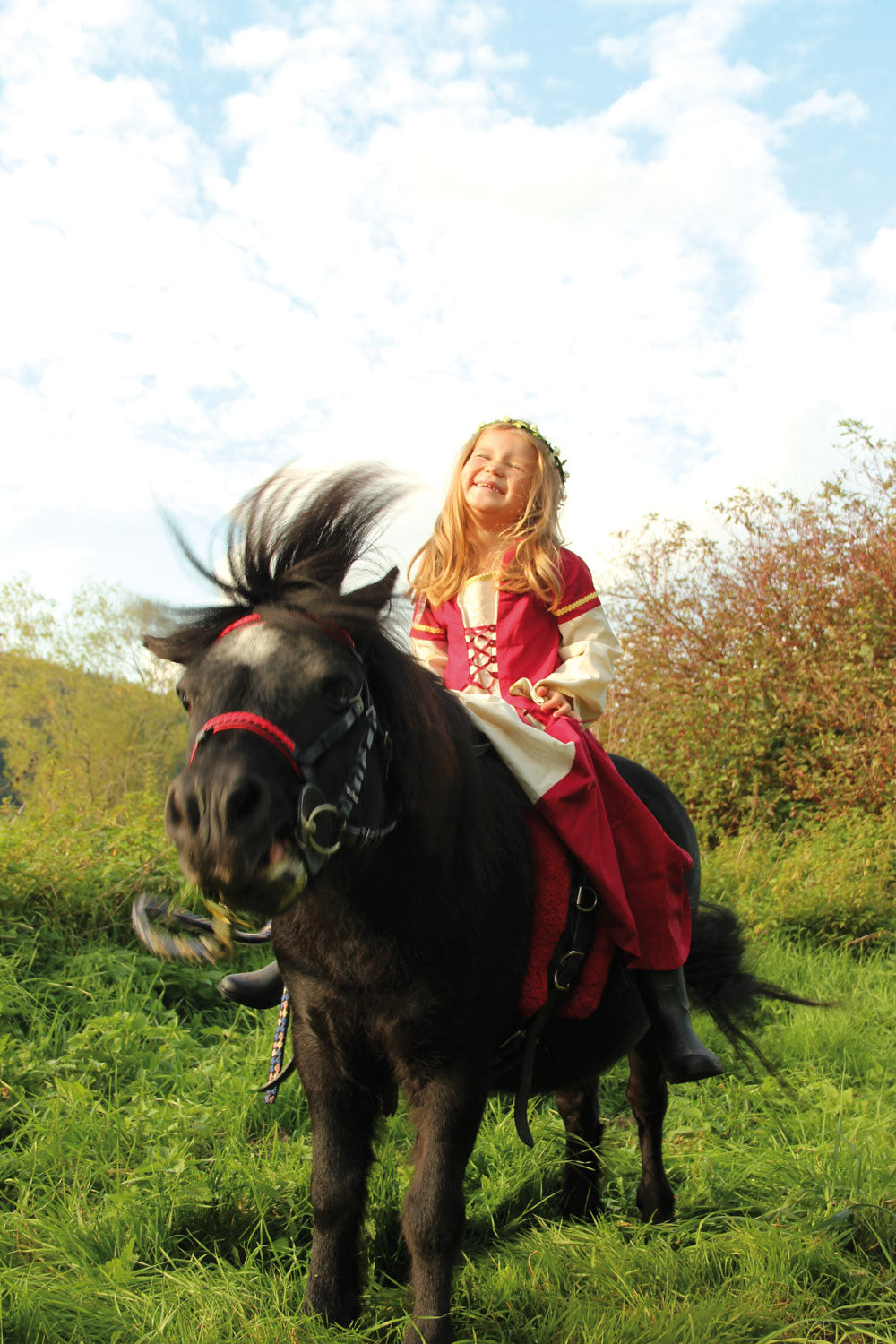 Child wearing the Vah Red Little Marian costume Dress with a cream coloured floral crown whilst riding a pony