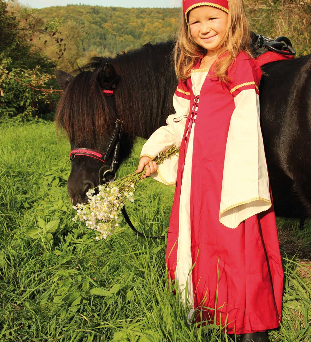 Child wearing the Vah Red Little Marian costume Dress with a matching headpiece holding a bunch of wild daisies. The child is standing next to a pony