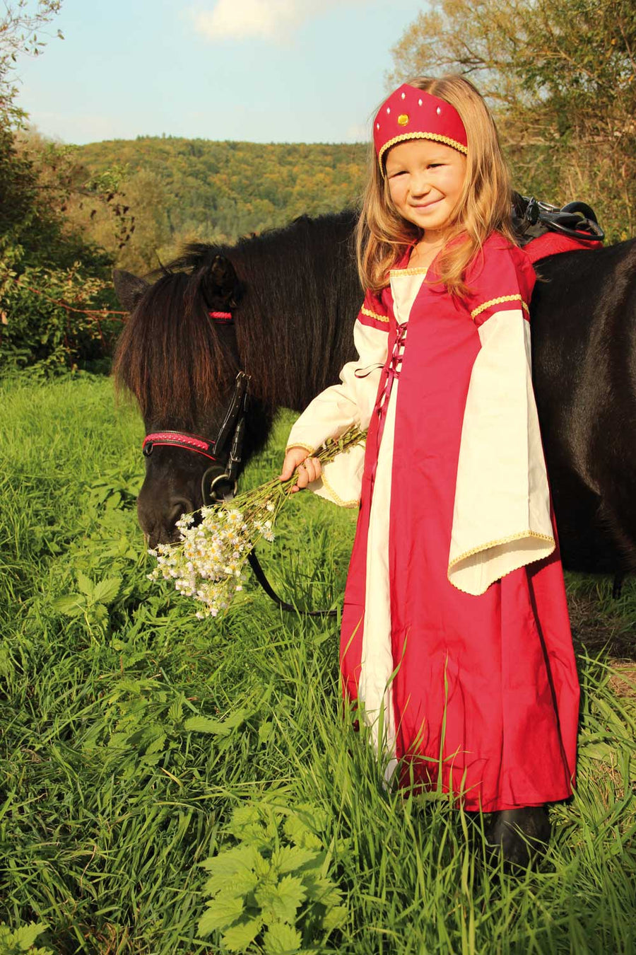 Child wearing the Vah Red Little Marian costume Dress with a matching headpiece holding a bunch of wild daisies. The child is standing next to a pony