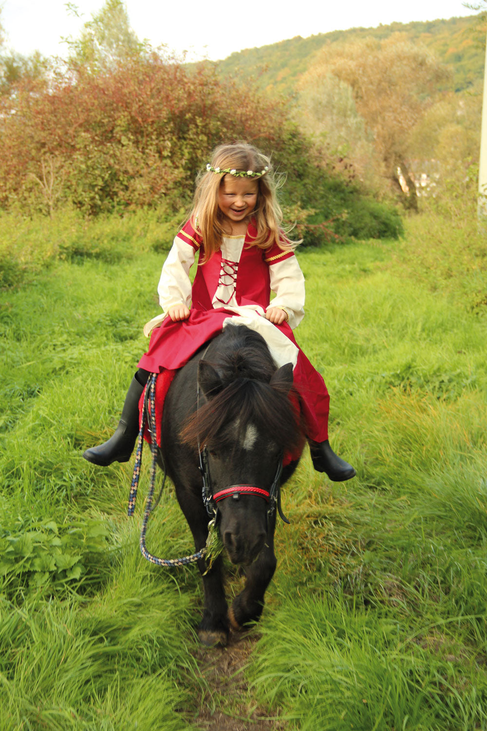 Child wearing the Vah Red Little Marian costume Dress with a cream coloured floral crown whilst riding a pony