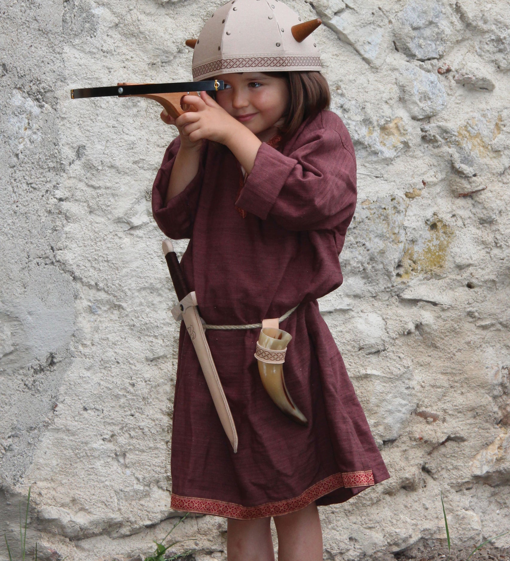 A child in full Viking costume including the Vah Viking Tunic, helmet and drinking horn. The child is playing with a cross bow.