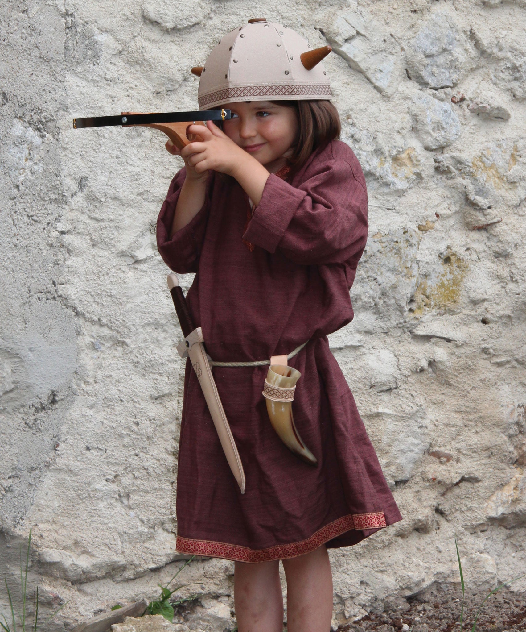 A child in full Viking costume including the Vah Viking Tunic, helmet and drinking horn. The child is playing with a cross bow.