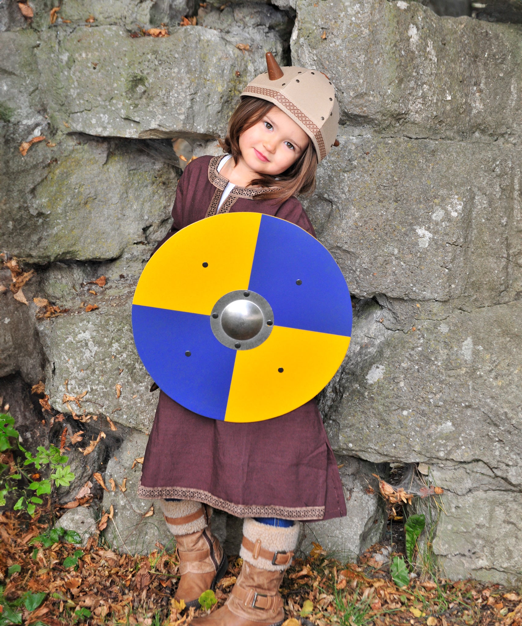 A child wearing the Vah Viking Tunic costume with a Viking helmet holding up a round toy shield.
