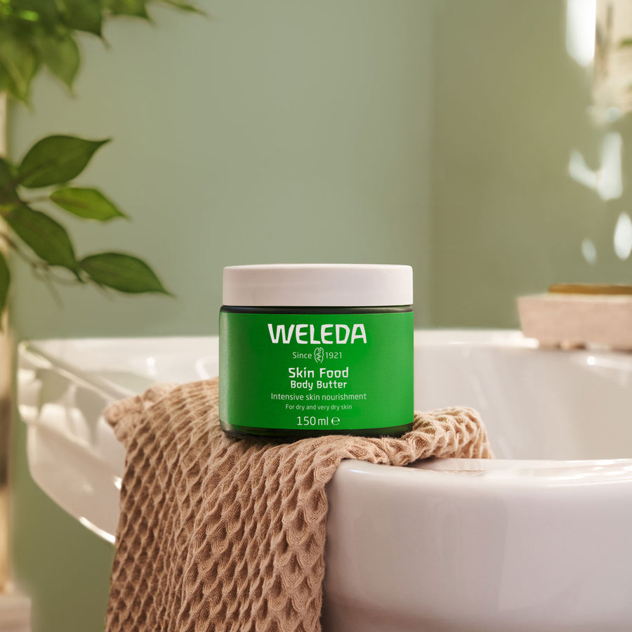 Weleda Skin Food Body Butter pot placed on a dusk pink coloured towel on the side of a bath. A green leafed plant can be seen in the background