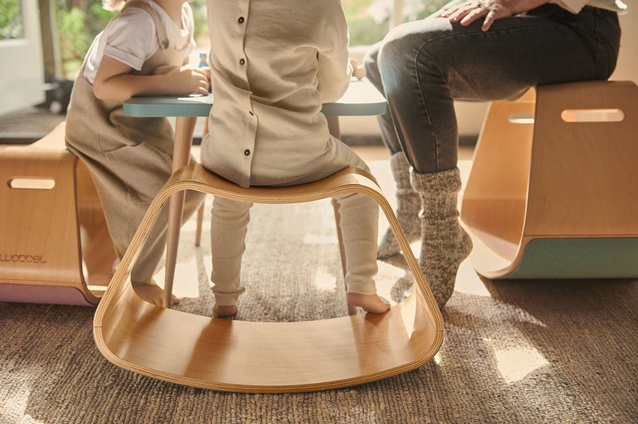 Close up of 3 child sat at a table on a Wobbel curved wooden Up balance box on a grey carpet