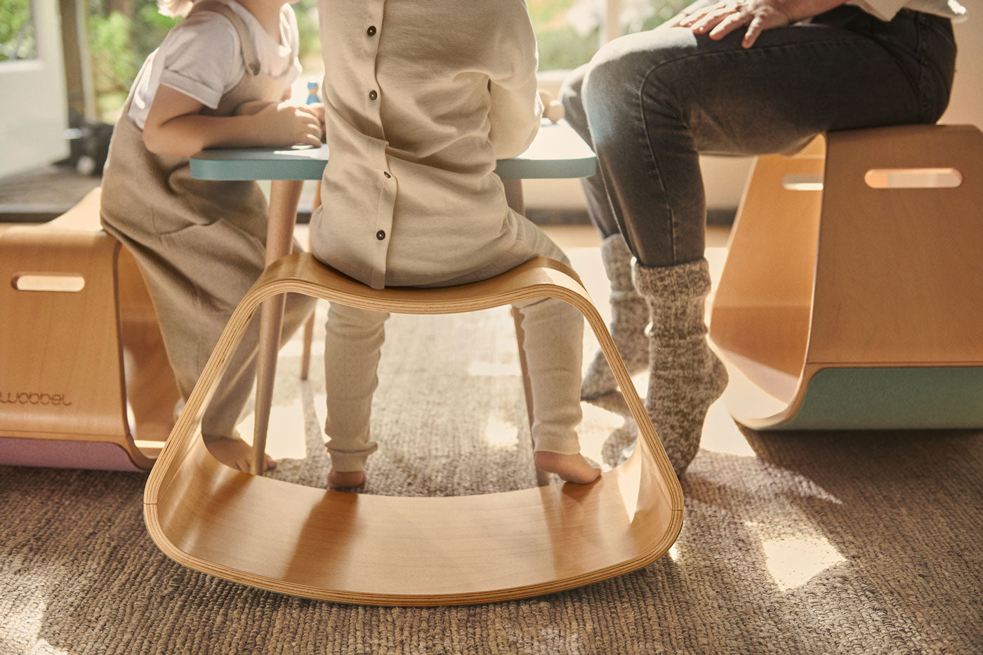 Close up of 3 child sat at a table on a Wobbel curved wooden Up balance box on a grey carpet