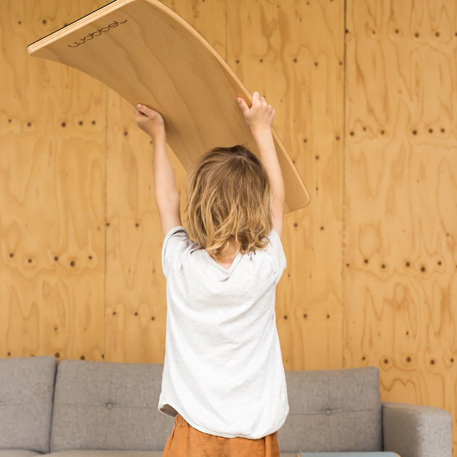 Child holding Wobbel Starter Beech Wood wobble board above his head in front of a wooden wall