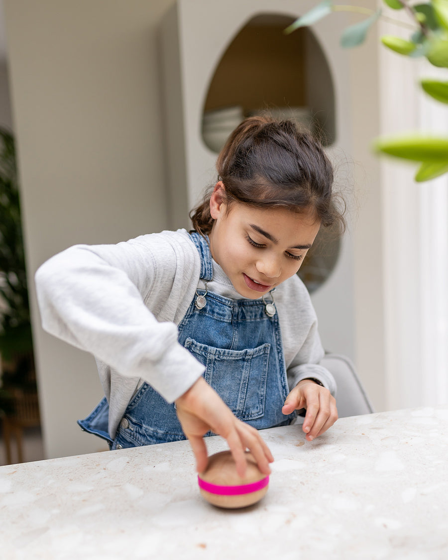 A child playing and spinning a Wooden Wobbel Candy Big Macaron on a  table. Made from Beech wood with a pink "filling"