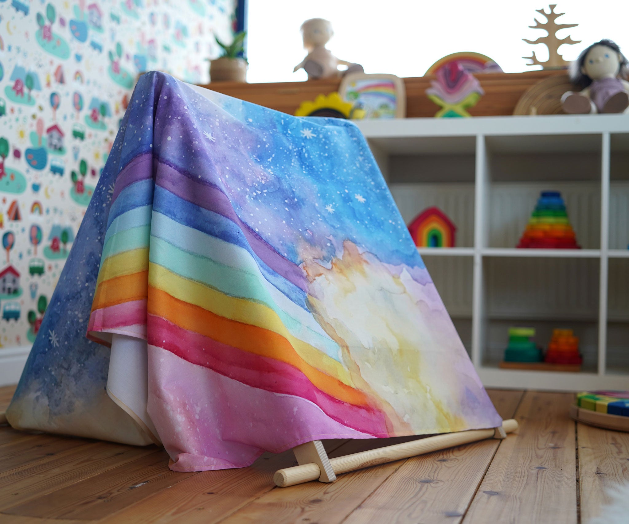 A giant Wonderie play cloth with the magical sky design placed over a Triclimb climbing frame to create a den. Shelves filled with toys can be seen in the background. 