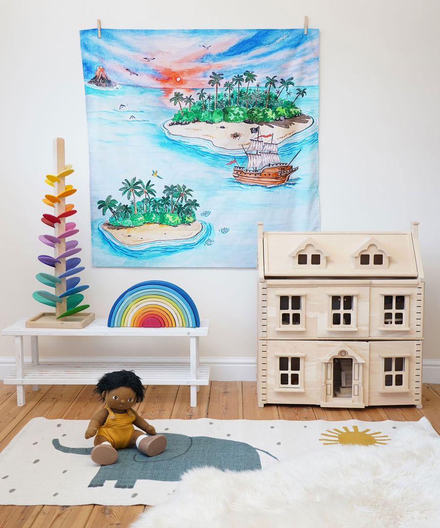 The Wonderie The Lost Island design play cloth hung up on the wall in a playroom. The PlanToys Victorian dolls house, Magic Wood marble tree and the Gwawr rainbow are placed in front. An Olli Ella doll can be seen sitting on the floor at the front on a rug. 