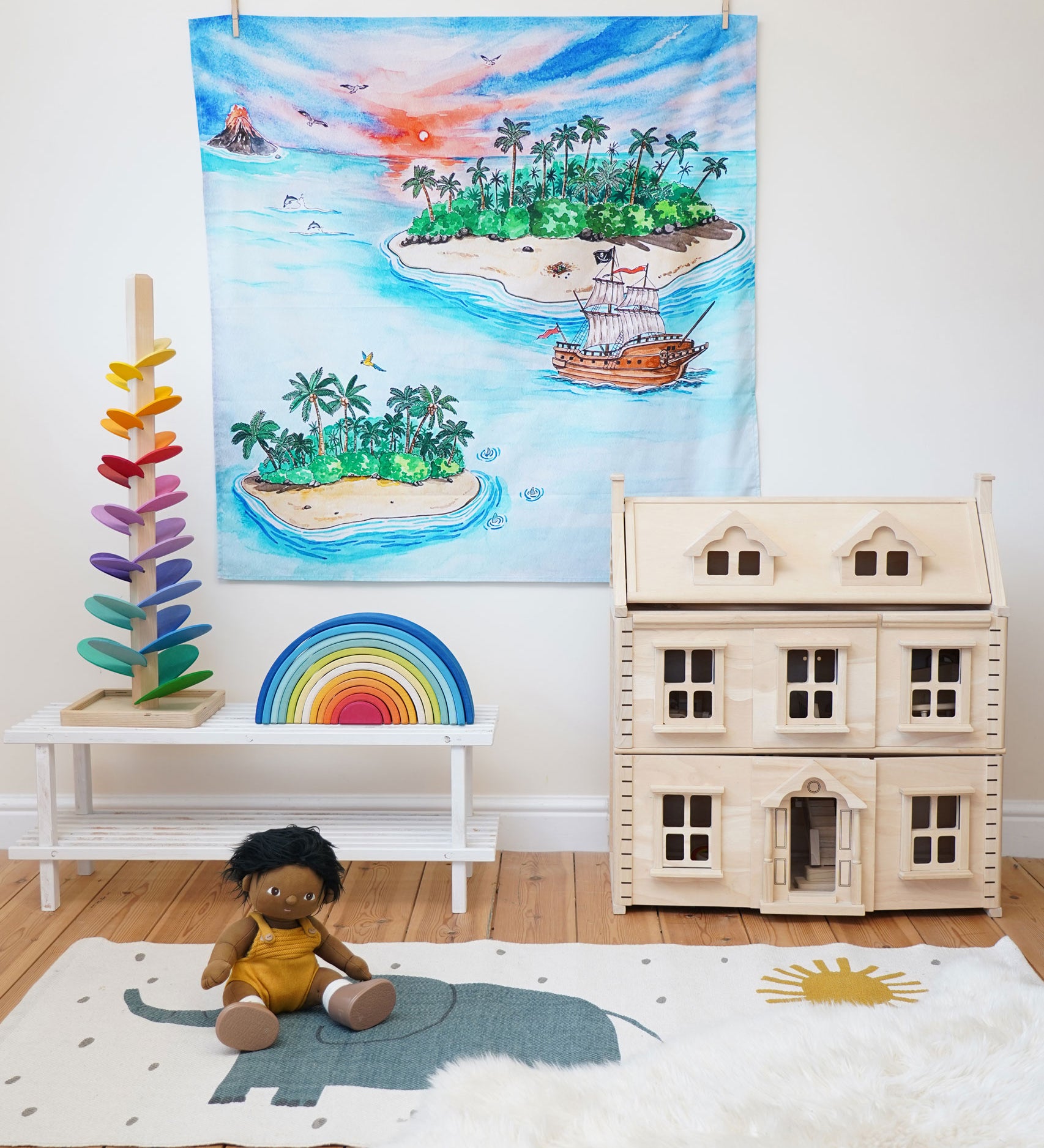 The Wonderie The Lost Island design play cloth hung up on the wall in a playroom. The PlanToys Victorian dolls house, Magic Wood marble tree and the Gwawr rainbow are placed in front. An Olli Ella doll can be seen sitting on the floor at the front on a rug. 