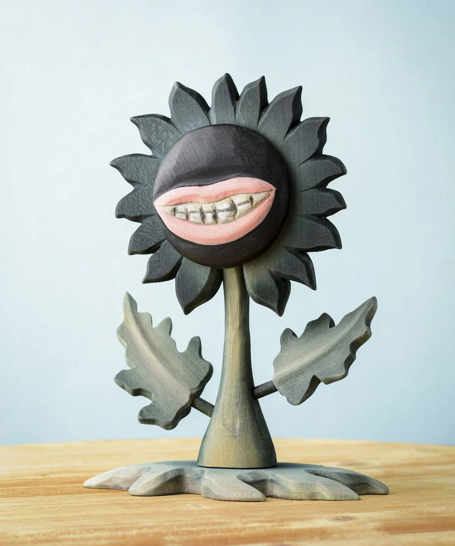 Bumbu Toys black wooden sunflower figure with a mouth with light pink lips and white teeth carved into it for a creepy Halloween vibe. The toy sunflower figure is placed on a wooden surface. 