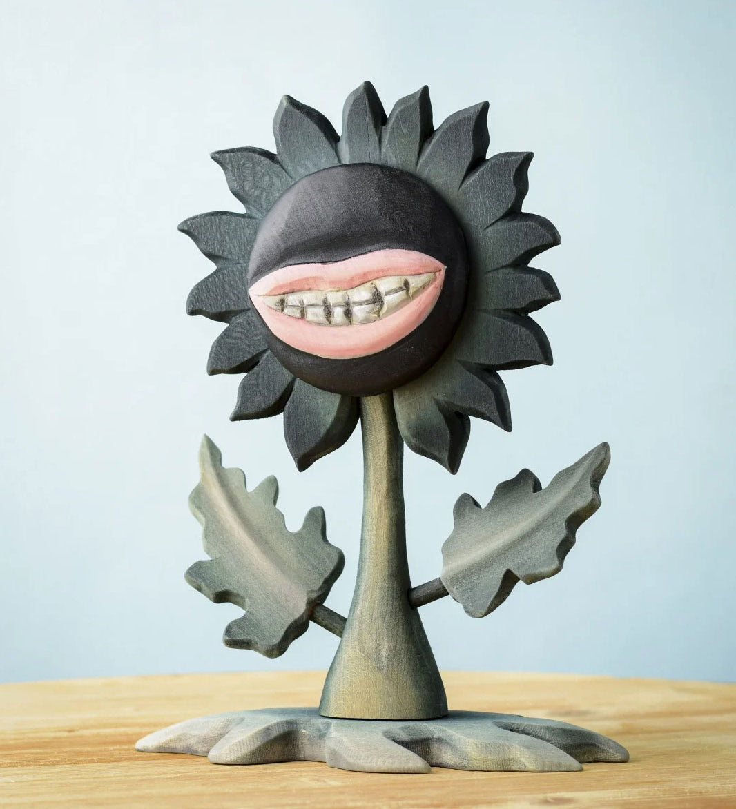 Bumbu Toys black wooden sunflower figure with a mouth with light pink lips and white teeth carved into it for a creepy Halloween vibe. The toy sunflower figure is placed on a wooden surface. 