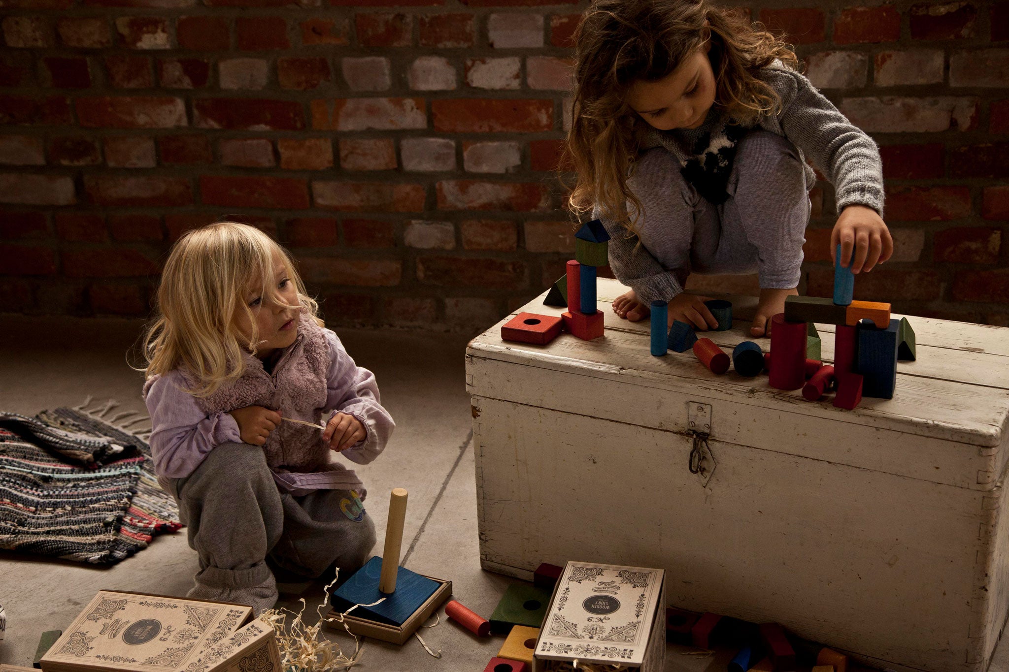 2 children playing with the Wooden Story 30 Rainbow Blocks in Tray. One child is sat on the floor the other is sat on top of a painted wooden chest. 