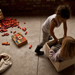 Wooden Story Rainbow Blocks in Tray on the floor with children playing next to them. A wooden box can be seen in the background surrounded by yellow and orange blocks. 