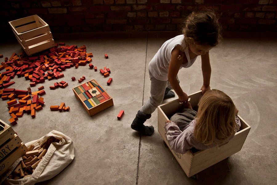 Wooden Story Rainbow Blocks in Tray on the floor with children playing next to them. A wooden box can be seen in the background surrounded by yellow and orange blocks. 