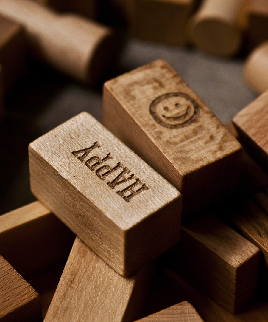 A close up of the blocks from the Wooden Story 72 XL Peace & Love Blocks set. There are two block on the top with etched details, one has a simple smiley face the other has the word happy written on it in capital letters. 