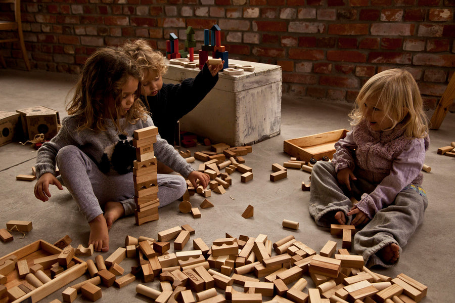 Children sitting on a floor playing with blocks from various Wooden Story block set. The three children are surrounded by blocks of different sizes and shapes. 