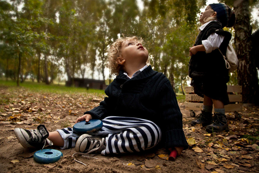 A child playing with two blue pieces from the Wooden Story Rainbow Stacker. The child is sitting on a muddy floor outdoors surrounded by fallen leaves and trees. 