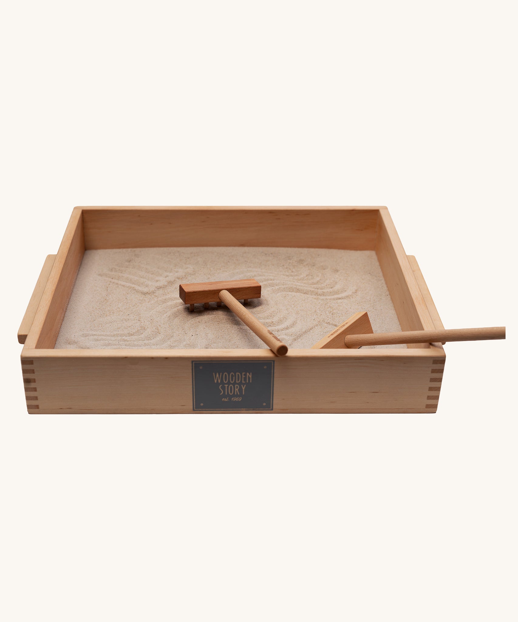 Wooden Story Montessori Sand Tray and Accessories shown on a plain background. The sand is in the tray with the tools. The tray is placed at an angle and the printed logo can be seen on the side. 