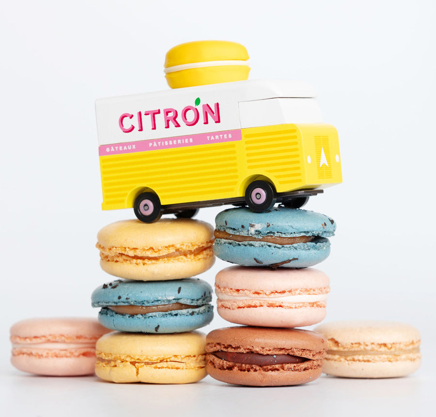 Picture of the Citron Candylab Candyvan on top of a pile of macarons. It is a yellow and white van with a yellow Lemon Macaron on the roof.